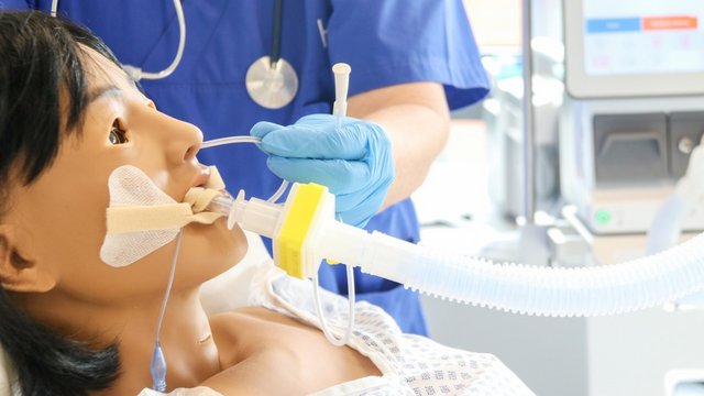 Simulationspuppe mit Beatmungstubus, daneben eine Person im blauen Kasack vor einem Klinikmonitor